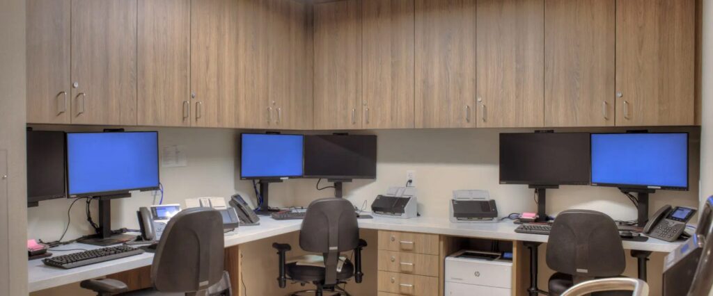 A modern office workspace with four monitors, arranged in pairs on desks facing each other. Each station has a computer, phone, and office chair. The walls are lined with sophisticated wooden cabinetry, and there is additional office equipment on the desks. The scene is well-lit.