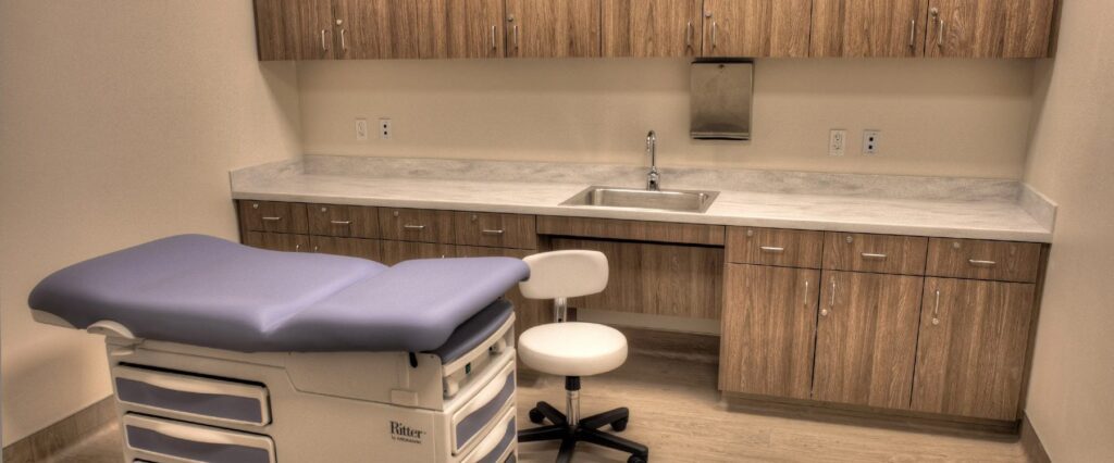 A modern medical examination room featuring a cushioned exam table with drawers underneath, an adjustable white stool, and a countertop with a sink. Wooden cabinetry lines the back wall above and below the countertop.